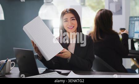 Designer analyzing 3D architectural model with printed maquette in shared workspace. Asian architect specializing in industrial development, using manufacturing drafts. Handheld shot. Stock Photo
