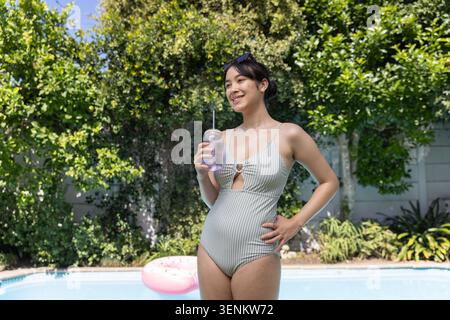 Asian woman standing on pool deck, holding tumbler beside pink inflatable ring floating in pool Stock Photo