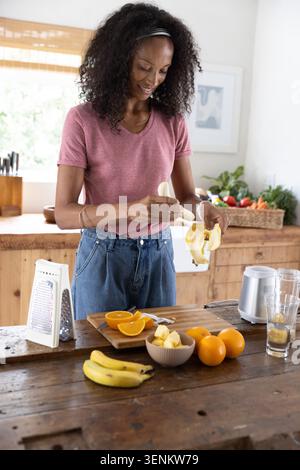 African American woman peeling banana at wooden island with sliced oranges and pineapple in kitchen Stock Photo