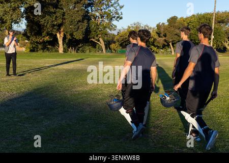 Four male cricket players in same shirts walking on field carrying helmets, bat, pads, copy space Stock Photo