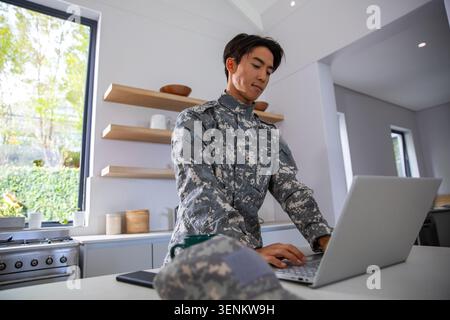 Asian man in camouflage uniform standing at kitchen island, using silver laptop with cap nearby Stock Photo