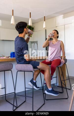 Diverse couple sitting on padded stools at wooden island in activewear, drinking from clear glasses Stock Photo
