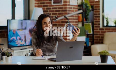 Laughing happy Indian woman taking selfie with mobile phone while smiling at home office desk. Person in nice stylish modern house capturing photo using smartphone camera, camera B. Stock Photo