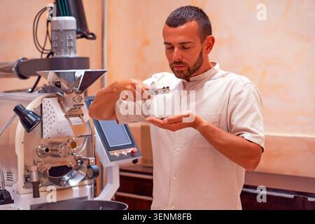 Bearded worker near coffee roaster controlling level of grain roasting, sniffs a sample portion of roasted beans. Stock Photo