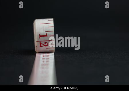 Selective focus on a white measuring tape roll with red numbers extending on a dark surface Stock Photo