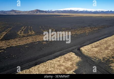 Drone aerial landscape of Black sand desert in iceland with sparse grass growing in patterns, a road, and snow capped mountains in the background. Stock Photo