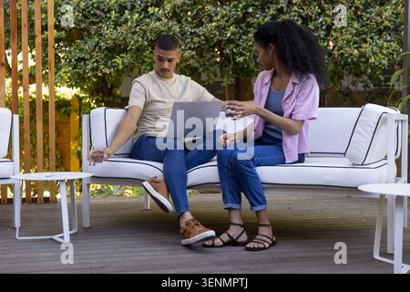 Diverse couple sitting on white outdoor sofa on backyard deck, passing glass and using laptop Stock Photo