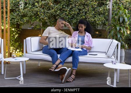 Diverse coworkers collaborating on white outdoor sofa on wooden deck with laptop, papers, pen Stock Photo