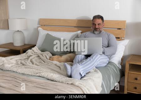Senior man sitting by headboard on bed in pajamas using laptop with napping dog, copy space Stock Photo