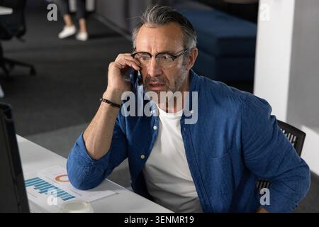 Middle-aged man holding smartphone and reviewing charts at office desk wearing blue shirt Stock Photo