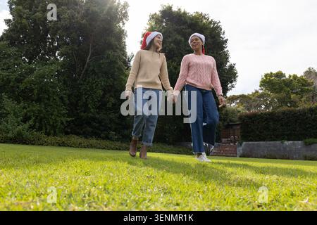 Asian female friends walking hand-in-hand on backyard lawn, wearing Santa hats and sweaters Stock Photo