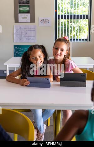Diverse school-age girls sitting together at classroom table using tablets in folio covers Stock Photo