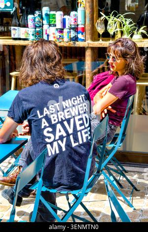 Young couple talking in café with man wearing T-shirt with "Last Night a Brewer Saved my Life" humour message - Tours, Indre-et-Loire (37), France. Stock Photo
