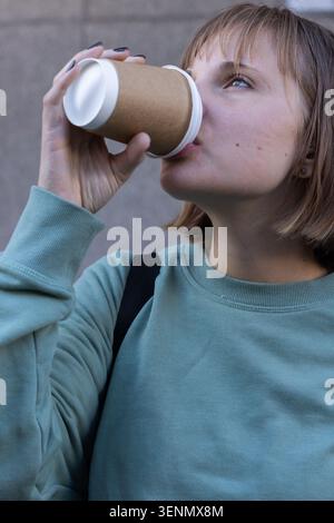 Woman tilting head back, drinking from disposable cup near stone wall, showing teal top Stock Photo