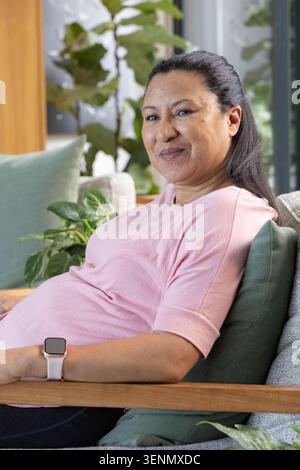Mature African American woman sitting on sofa with green pillows, wearing smartwatch near window Stock Photo