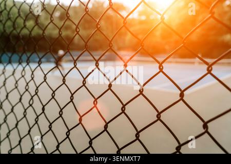 A shallow depth-of-field shot focusing on the geometric pattern of a black chain-link fence, with a soft-blurred tennis court and a hint of a player visible in the golden glow of the setting sun, creating a contrast between structure and atmosphere. Stock Photo
