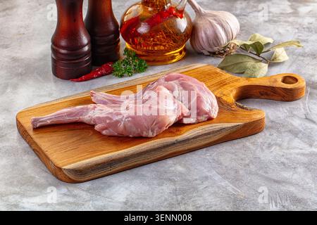 Fresh raw rabbit legs on wooden board with garlic, oil, pepper mills, and herbs on stone surface, ready for cooking preparation Stock Photo