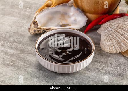 Dark brown Japanese oyster sauce dressing dip in white ceramic bowl with oyster shell, chili pepper, wooden spoon on grey stone background. Asian cuis Stock Photo