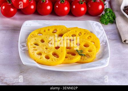 Yellow marinated pickled lotus root slices on white square plate with cherry tomatoes and parsley on light gray background Stock Photo