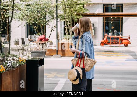 Woman standing outdoors sips beverage while holding smartphone and carrying woven tote bag. Sustainable living, urban lifestyle, eco friendly choices. Stock Photo