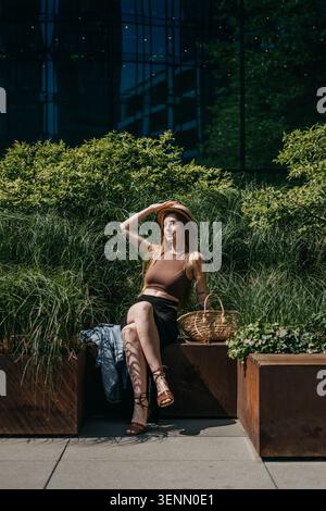 Woman sits on outdoor bench surrounded by green vegetation holding straw hat. Mitigating urban heat, cool city infrastructure design, smart urban Stock Photo