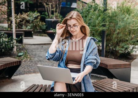 Woman smiles while working on laptop on public bench outdoors. Women in technology, successful young woman, self-employed female. Stock Photo