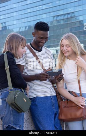 Diverse friends standing at plaza on stone ledge, sharing smartphone near reflective glass Stock Photo