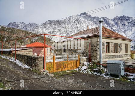 Two traditional stone and stucco houses with vibrant red tiled roofs and a rusted orange gate stand on a dirt path with patches of snow. In the backgr Stock Photo