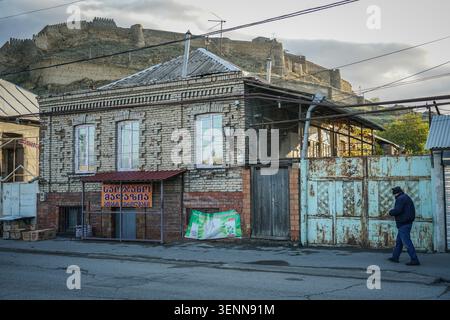 A man walks along a street past an old brick building with a Georgian script sign under a red awning, opposite a rusty metal gate. In the background, Stock Photo