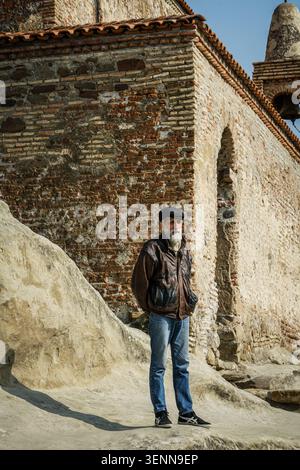 An elderly man with a long white beard, wearing a brown leather jacket and a flat cap, stands on the carved rock ground of Uplistsikhe cave town. In t Stock Photo