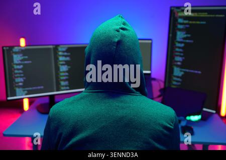 Hooded hacker coding at dual monitors in neon lit room Stock Photo