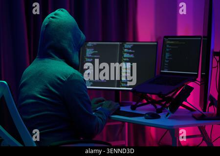 Hooded Hacker Coding at Dual Monitors in Dark Neon Room Stock Photo