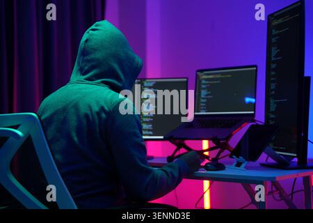 Hooded Hacker Working at Multiple Monitors in Dark Room Stock Photo