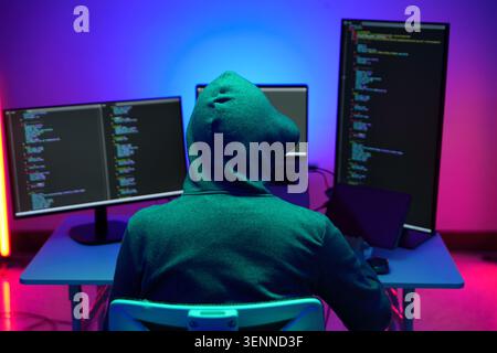 Hacker in Hoodie Working at Multiple Monitors in Neon Light Stock Photo