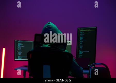 Hacker in Hoodie Working at Dual Monitors in Dark Neon Room Stock Photo