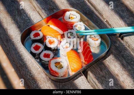 a close-up of assorted sushi and nigiri arranged in a stainless steel lunch box on a weathered wooden bench, with striped shadows crossing the surface Stock Photo
