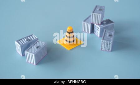 Yellow traffic cone placed between groups of miniature shipping containers on a light blue background indicating logistics delays or custom inspection Stock Photo