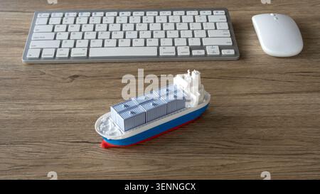 Toy cargo vessel loaded with shipping containers placed in front of a computer keyboard and mouse on a wooden workspace. Stock Photo
