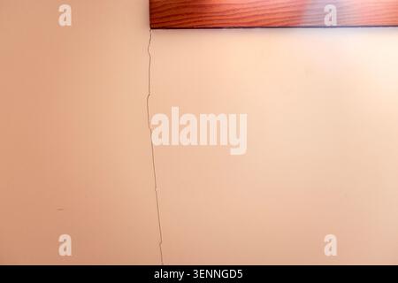 Crack in beige wall beneath wooden trim, indicating potential structural damage in a residential space, illuminated by natural light from nearby windo Stock Photo