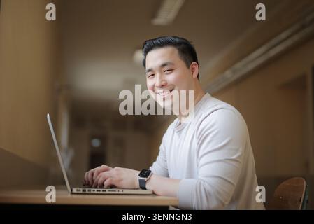 Smiling Asian man looks at camera while using laptop Stock Photo