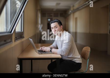 Young Asian man works on laptop in school hallway Stock Photo