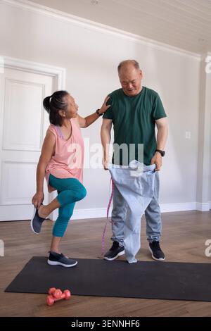 Asian couple balancing on mat at home, woman touching shoulder while man holding jeans with tape Stock Photo