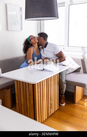 Diverse couple kissing and holding hands at home dining nook with coffee mug notebook. Affection, romance, partners, intimate, cozy, bright, daylight Stock Photo