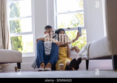 Couple sitting on floor by large windows taking selfie with smartphone and to-go cups nearby Stock Photo