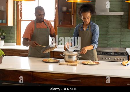 Diverse couple wearing aprons, preparing and plating pasta on wide white island using colander Stock Photo