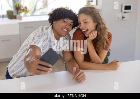 Couple posing and taking selfie with phone while leaning on white countertop in kitchen Stock Photo