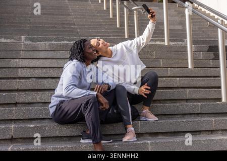 Diverse female friends sitting on concrete steps taking selfie with phone, metal rails and sneakers Stock Photo