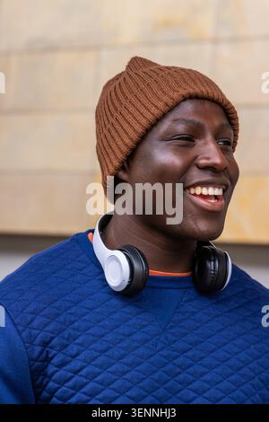 African American man smiling, looking right by wall, wearing beanie, quilted sweatshirt, headphones Stock Photo