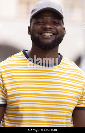 African man smiling with trimmed beard, wearing gray cap and yellow-white striped tee in plaza Stock Photo
