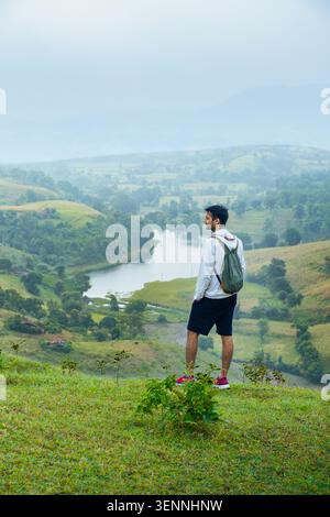 A young man stands on a grassy hill, admiring a serene lake surrounded by lush green landscapes in Rajasthan. Stock Photo